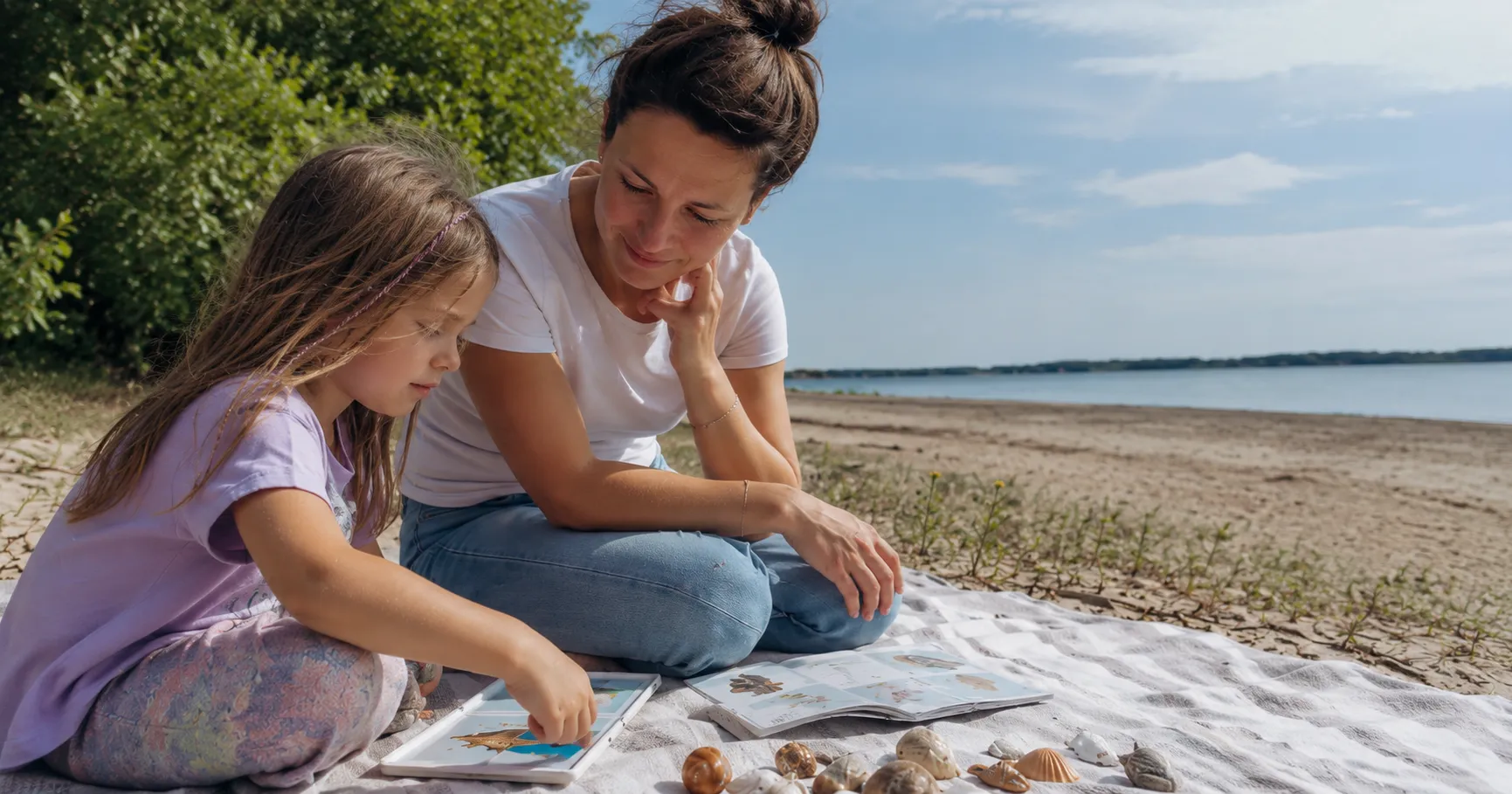 Mother and daughter naming shells and looking at a picture book on the beach – summer activities that support a child’s speech